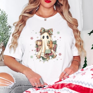 May include: A white short-sleeve t-shirt featuring a graphic of a ghost wearing a plaid bow, holding a mug and a gingerbread cookie. The ghost is wrapped in a plaid blanket and surrounded by small snowflakes, red dots, and a string of Christmas lights.