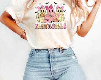 May include: A cream-colored t-shirt featuring a graphic of three ghosts wearing pink bows, holding a concha, a coffee cup, and a croissant, with small pumpkins and bats. The text PANTASMAS is printed in a retro, multi-colored font below the ghosts.