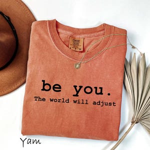 K&ouml;nnte beinhalten: Ein korallenfarbenes T-Shirt mit den Worten "be you. The world will adjust" in Schwarz gedruckt. Eine goldene Halskette mit Anh&auml;nger liegt auf dem Shirt. Ein brauner Hut und getrocknete Palmbl&auml;tter sind ebenfalls im Bild.