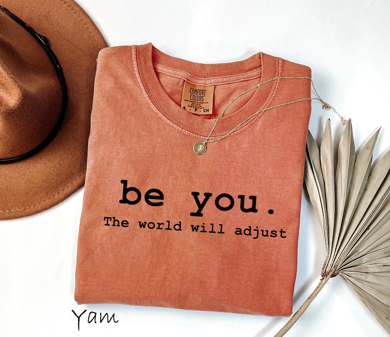 K&ouml;nnte beinhalten: Ein korallenfarbenes T-Shirt mit den Worten "be you. The world will adjust" in Schwarz gedruckt. Eine goldene Halskette mit Anh&auml;nger liegt auf dem Shirt. Ein brauner Hut und getrocknete Palmbl&auml;tter sind ebenfalls im Bild.