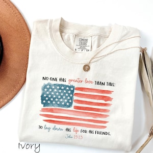 May include: Ivory-colored t-shirt with a watercolor American flag design. The flag features a blue square with white stars and red stripes. Text reads "NO ONE HAS greater love THAN THIS: To lay down HIS life FOR HIS FRIENDS. John 15:13."