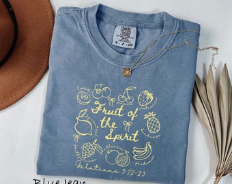 May include: A blue jean colored t-shirt with a crew neckline and a Comfort Colors brand label. The front features a yellow graphic print of various fruits and the text "Fruit of the Spirit" along with "Galatians 5:22-23" and small bows.