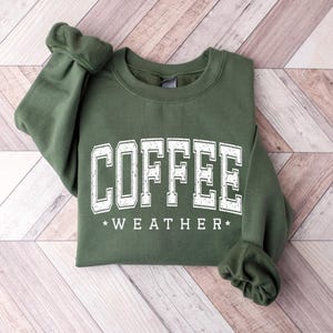 May include: An olive green crewneck sweatshirt featuring the word COFFEE in large, distressed white block letters, with the word WEATHER in smaller white letters underneath, flanked by two small white stars on either side of the text.