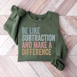 May include: An olive green crewneck sweatshirt featuring the text BE LIKE SUBTRACTION AND MAKE A DIFFERENCE printed in a distressed, multi-colored font. The top line is light blue, the middle line is pink, and the bottom line is yellow-gold.