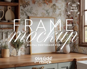 May include: A digital mockup of a framed picture in a kitchen setting. The frame is on a wooden countertop with a floral wallpaper background. The text "FRAME MOCKUP" is written in a stylized font above the frame. The text "DIGITAL PHOTOSHOP FILE" is written below the frame. The text "outside the mock" is written below the countertop.