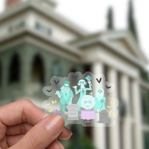 May include: A clear sticker featuring a group of cartoon ghosts from the Haunted Mansion attraction at Disneyland. The ghosts are dressed in formal attire and are holding a party. The sticker is being held in front of a blurred image of the Haunted Mansion.