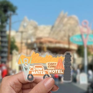 Puede incluir: Una pegatina transparente con un dise&ntilde;o estilizado del Cozy Cone Motel de la pel&iacute;cula Cars. La pegatina incluye el texto "Tow Mater Motel" y "Cozy Cone Motel" en naranja y blanco, con una pila de neum&aacute;ticos y un cono.