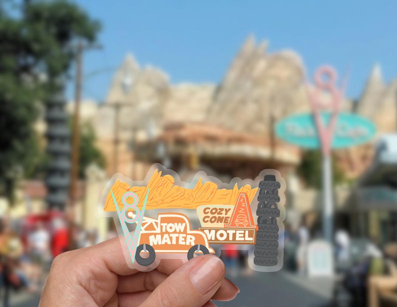 Puede incluir: Una pegatina transparente con un dise&ntilde;o estilizado del Cozy Cone Motel de la pel&iacute;cula Cars. La pegatina incluye el texto "Tow Mater Motel" y "Cozy Cone Motel" en naranja y blanco, con una pila de neum&aacute;ticos y un cono.