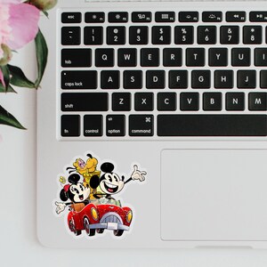 May include: A sticker featuring Mickey Mouse, Minnie Mouse, and Pluto driving a red car. The sticker is on a silver laptop.