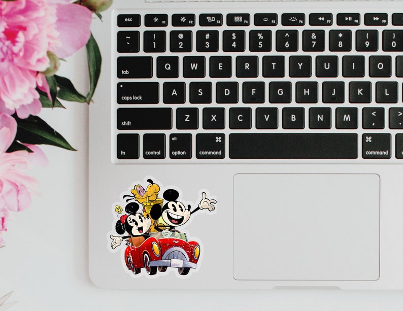 May include: A sticker featuring Mickey Mouse, Minnie Mouse, and Pluto driving a red car. The sticker is on a silver laptop.
