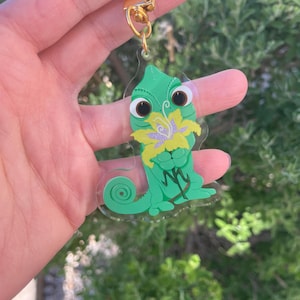 May include: A green chameleon keychain with a gold-colored clasp. The chameleon has large brown eyes and is holding a yellow and purple flower. The keychain is made of clear acrylic.