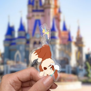 May include: A clear sticker featuring a cartoon dog with brown fur and a blue collar. The dog is looking up with a surprised expression. A small fairy with green wings and a yellow dress is flying above the dog, sprinkling glitter.