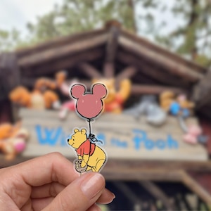 May include: A yellow Winnie the Pooh character pin holding a pink Mickey Mouse balloon. The pin features a black outline and a red shirt. The background is blurred, but appears to be a theme park.