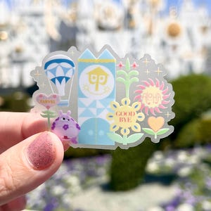 May include: A clear sticker featuring a whimsical design with a hot air balloon, a smiling clock tower, and a purple creature. The sticker includes the words "FARVEL", "ALOHA", and "GOOD BYE" in various fonts and colors. The background is blurred.