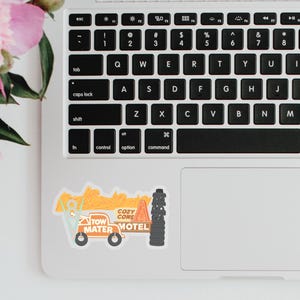 Puede incluir: Una pegatina con un cami&oacute;n de remolque de dibujos animados con el texto "Tow Mater" y "Cozy Cone Motel". La pegatina est&aacute; en un port&aacute;til junto a flores rosas. El cami&oacute;n de remolque es naranja, azul y negro.