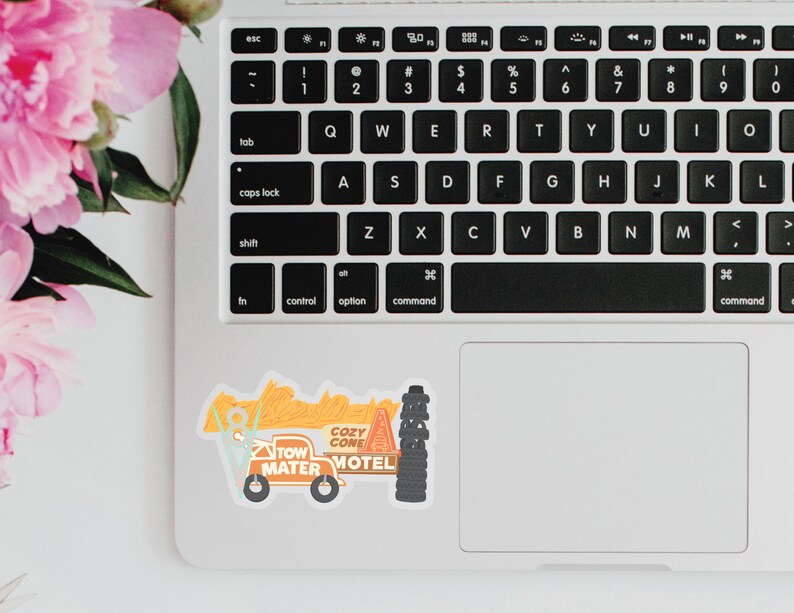 Puede incluir: Una pegatina con un cami&oacute;n de remolque de dibujos animados con el texto "Tow Mater" y "Cozy Cone Motel". La pegatina est&aacute; en un port&aacute;til junto a flores rosas. El cami&oacute;n de remolque es naranja, azul y negro.