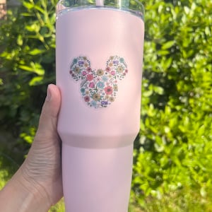 May include: A pink stainless steel tumbler with a floral Mickey Mouse design. The tumbler has a lid and a straw.