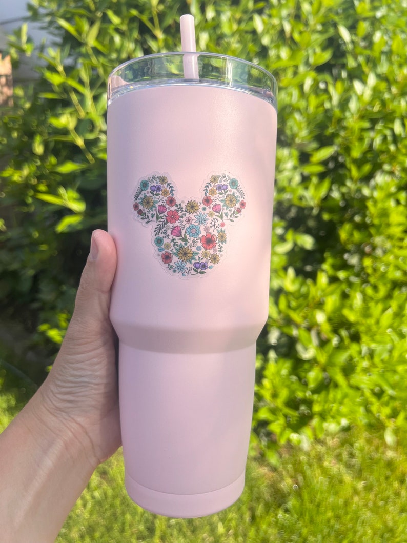 May include: A pink stainless steel tumbler with a floral Mickey Mouse design. The tumbler has a lid and a straw.