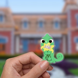 May include: A green chameleon sticker holding a yellow and purple flower. The sticker has large, expressive eyes and a curled tail. The background is blurred, showing a building and greenery.