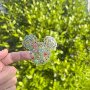 May include: A clear sticker with a floral design in the shape of Mickey Mouse's head. The sticker is on a finger against a blurred background of green foliage.