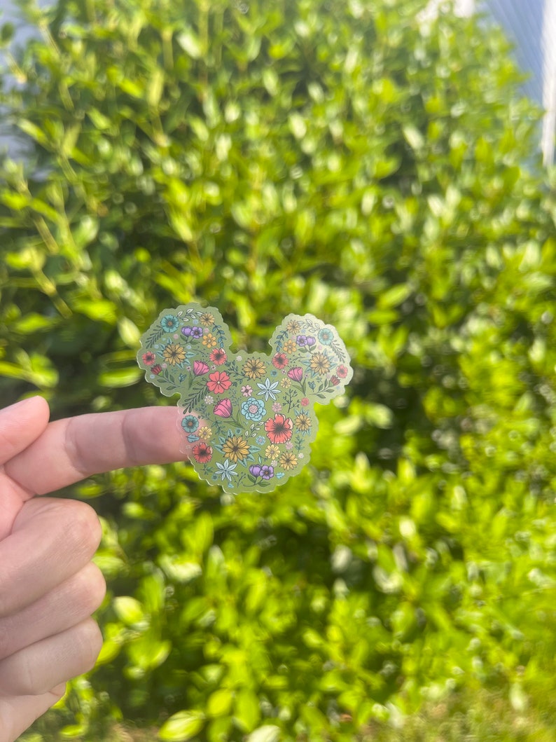 May include: A clear sticker with a floral design in the shape of Mickey Mouse's head. The sticker is on a finger against a blurred background of green foliage.