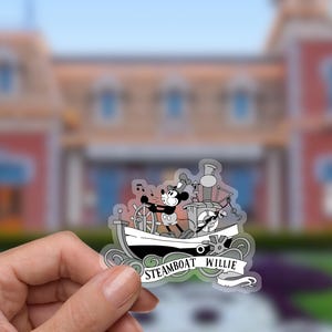 May include: A clear sticker featuring Mickey Mouse as Steamboat Willie, steering a steamboat. The black and white image includes musical notes and the text "Steamboat Willie" on a banner. The background is blurred.