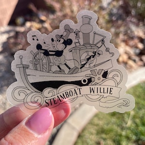 May include: A transparent sticker featuring a black and white illustration of Mickey Mouse at the helm of a steamboat. The text "Steamboat Willie" is displayed on a banner below the boat. Musical notes float above Mickey's head.