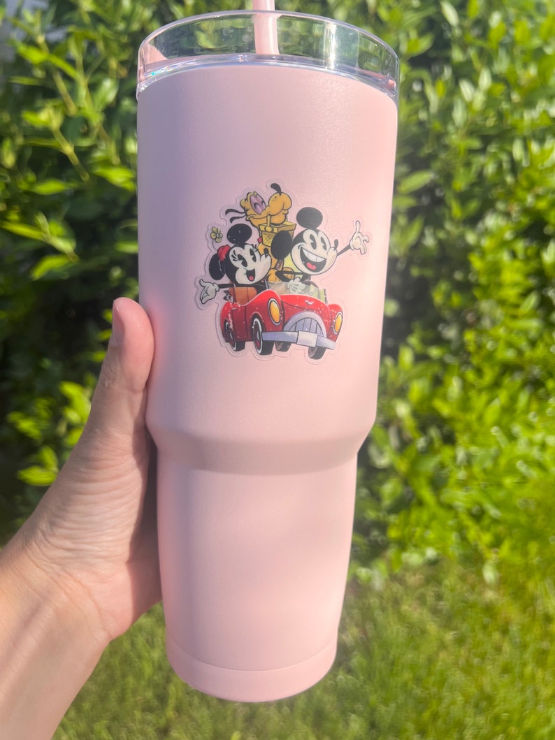 May include: A pink stainless steel tumbler with a Mickey Mouse, Minnie Mouse, and Pluto sticker. The sticker shows the characters riding in a red car.