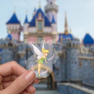 May include: A transparent sticker featuring Tinkerbell in a green dress with white wings and a blonde bun. The sticker has a glittery, sparkling design around the character. The background is a blurred image of a castle.