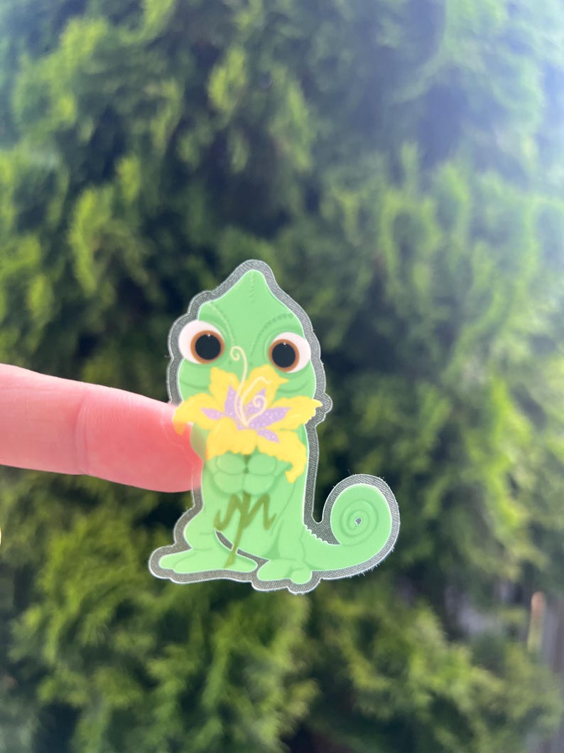 May include: A green chameleon sticker with large brown eyes and a yellow and purple flower. The sticker is held by a finger against a blurred green background.