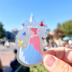 May include: A clear sticker featuring a princess in a pink and blue gown with birds flying around her. The sticker is held in a hand against a blurred background.