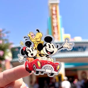 May include: A clear sticker featuring Mickey Mouse, Minnie Mouse, and Pluto in a red car. The sticker is held up in front of a blurred background of a building with the word "Disney" on it.