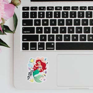 May include: A sticker featuring Ariel, the Little Mermaid, with her signature red hair and green tail. She is surrounded by colorful sea creatures and flowers.
