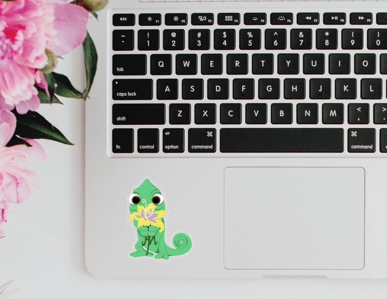 May include: A green chameleon sticker with a yellow flower is placed on a laptop. The laptop is white with a black keyboard. Pink flowers are visible in the background.