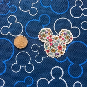 May include: A white sticker with a floral design in the shape of Mickey Mouse ears. The sticker is on a blue background with white Mickey Mouse ears.