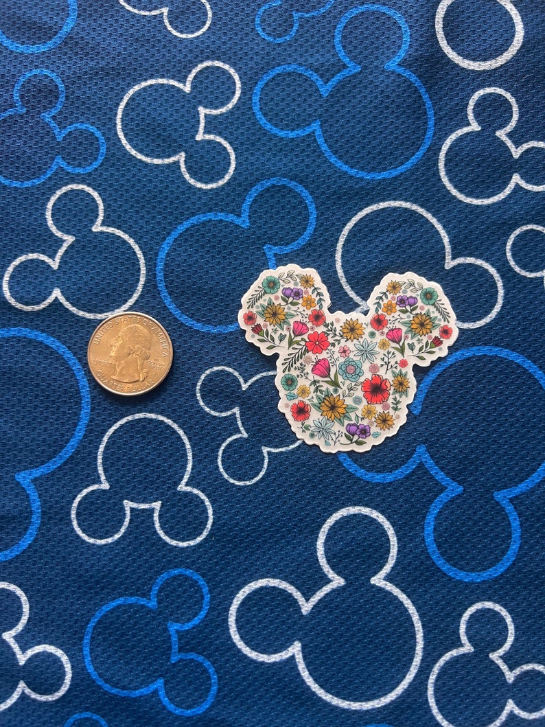 May include: A white sticker with a floral design in the shape of Mickey Mouse ears. The sticker is on a blue background with white Mickey Mouse ears.