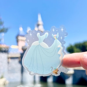 May include: A clear sticker featuring a blue Cinderella dress with a white bodice and a light blue cape. The sticker is placed on a finger in front of a blurred background of a castle.