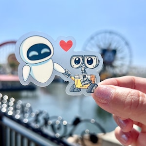 May include: A clear sticker with a white robot holding a red heart and a yellow robot with the word "WALL-E" on it.