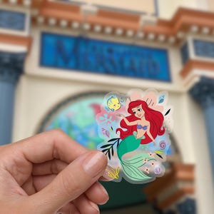 May include: A clear sticker featuring Ariel, the Little Mermaid, with a green tail and red hair. She is swimming with a yellow fish and surrounded by colorful sea life.