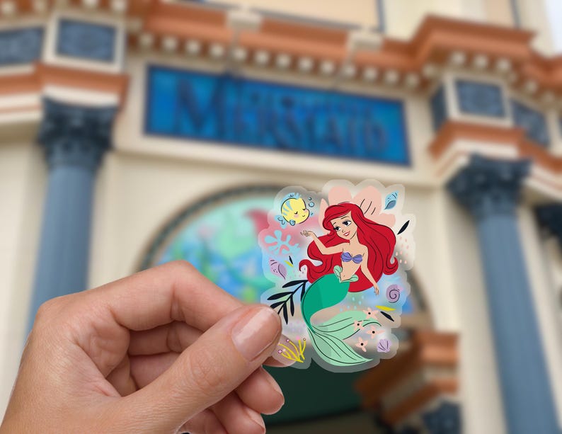 May include: A clear sticker featuring Ariel, the Little Mermaid, with a green tail and red hair. She is swimming with a yellow fish and surrounded by colorful sea life.