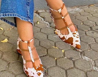 May include: Heels with a cowhide pattern in brown and white, featuring a pointed toe. The shoes have multiple straps and a zip. The shoes are worn with blue jeans.