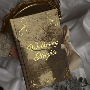 May include: A hardcover book titled "Wuthering Heights" by Emily Brontë. The book has a sepia-toned cover with gold lettering and decorative flourishes. A gold-colored tray and a satin ribbon are also visible.
