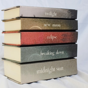 May include: A stack of five hardback books with the titles "twilight", "new moon", "eclipse", "breaking dawn", and "midnight sun". The books are in shades of grey, brown, and red, with white lettering.