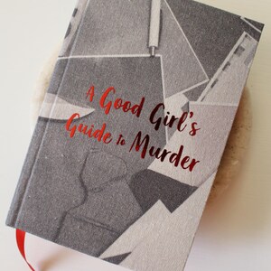 以下が含まれることがあります： 「A Good Girl's Guide to Murder」というタイトルのハードカバー本。グレーの幾何学模様の表紙と赤いリボンしおりが付いています。タイトルは赤いメタリック文字です。