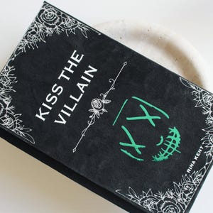 以下が含まれることがあります： 「Kiss the Villain」というタイトルの黒い本。白い文字とコーナーに花の模様があります。表紙には緑色のグラフィック要素と著者の名前が描かれています。本はニュートラルな背景に置かれています。