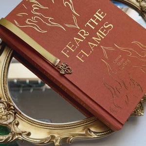 May include: A hardback book titled "FEAR THE FLAMES" in gold lettering, with flame illustrations. The book is rust-colored and has a gold ribbon bookmark with a dragon charm. The book rests on a gold-framed mirror.
