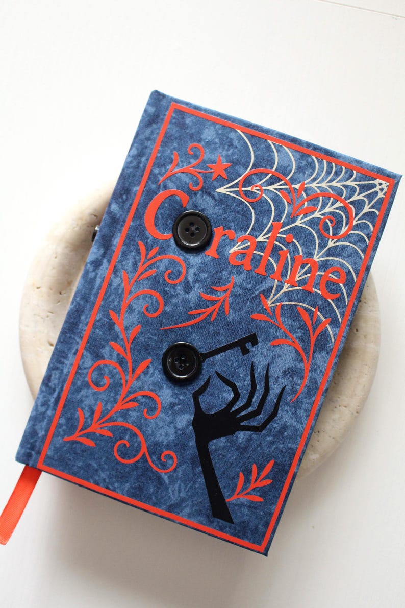 May include: A blue book with the title "Coraline" in red lettering. The cover features a spiderweb, key, and hand design. Two black buttons secure the cover. A red ribbon bookmark is visible.
