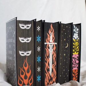 May include: A collection of six books with black edges featuring hand-painted designs. The first book cover reads A Court of Silver Flames. The page edges display white masks, orange flames, blue snowflakes, a white sword, gold stars, and flowers.