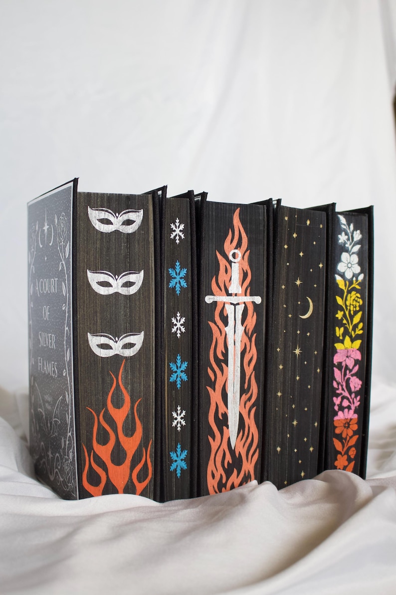 May include: A collection of six books with black edges featuring hand-painted designs. The first book cover reads A Court of Silver Flames. The page edges display white masks, orange flames, blue snowflakes, a white sword, gold stars, and flowers.