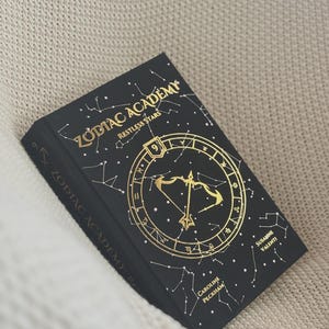 May include: A black hardback book titled Zodiac Academy: Restless Stars, featuring gold foil constellations and a central bow and arrow symbol. The cover displays the number 9 and the authors' names, Caroline Peckham and Susanne Valenti.